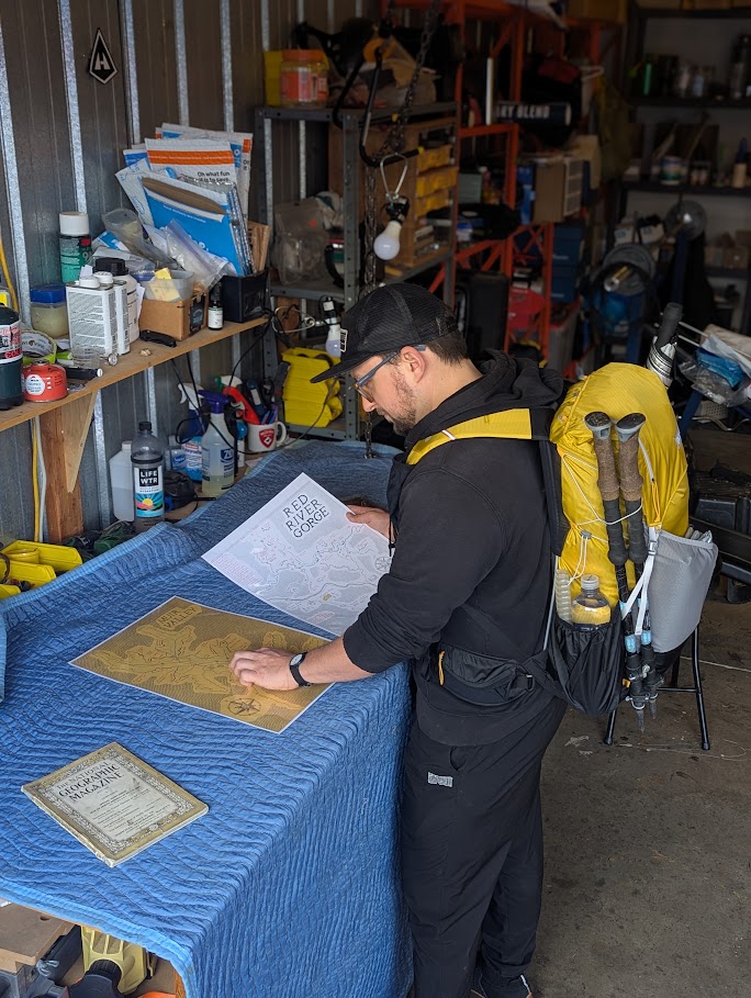 Justin Sovine in a garage wearing a loaded yellow backpack with trekking poles, studying a Red River Gorge map spread out on a workbench