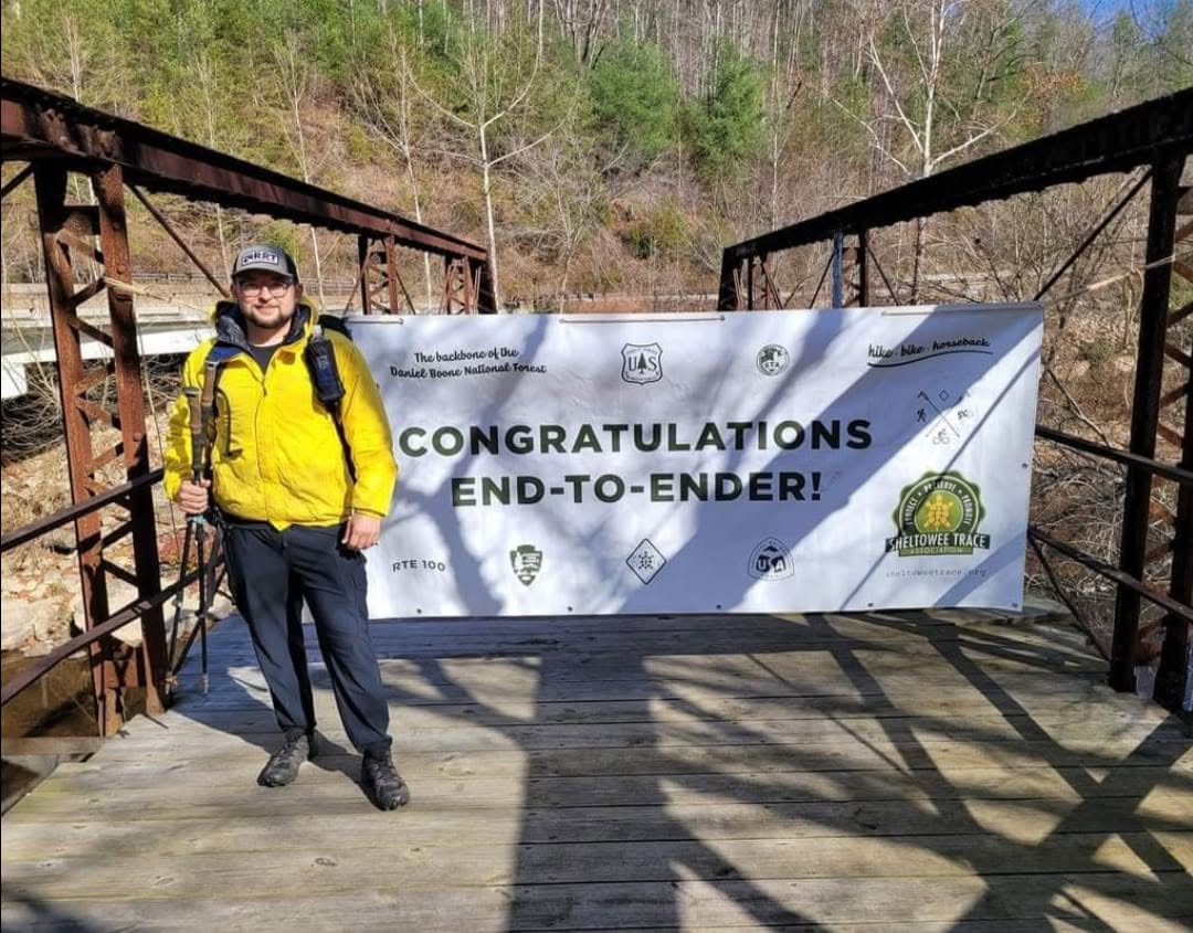 Justin Sovine on a bridge at the southern end of the Sheltowee Trace, next to a banner that reads “Congratulations End-to-Ender!”