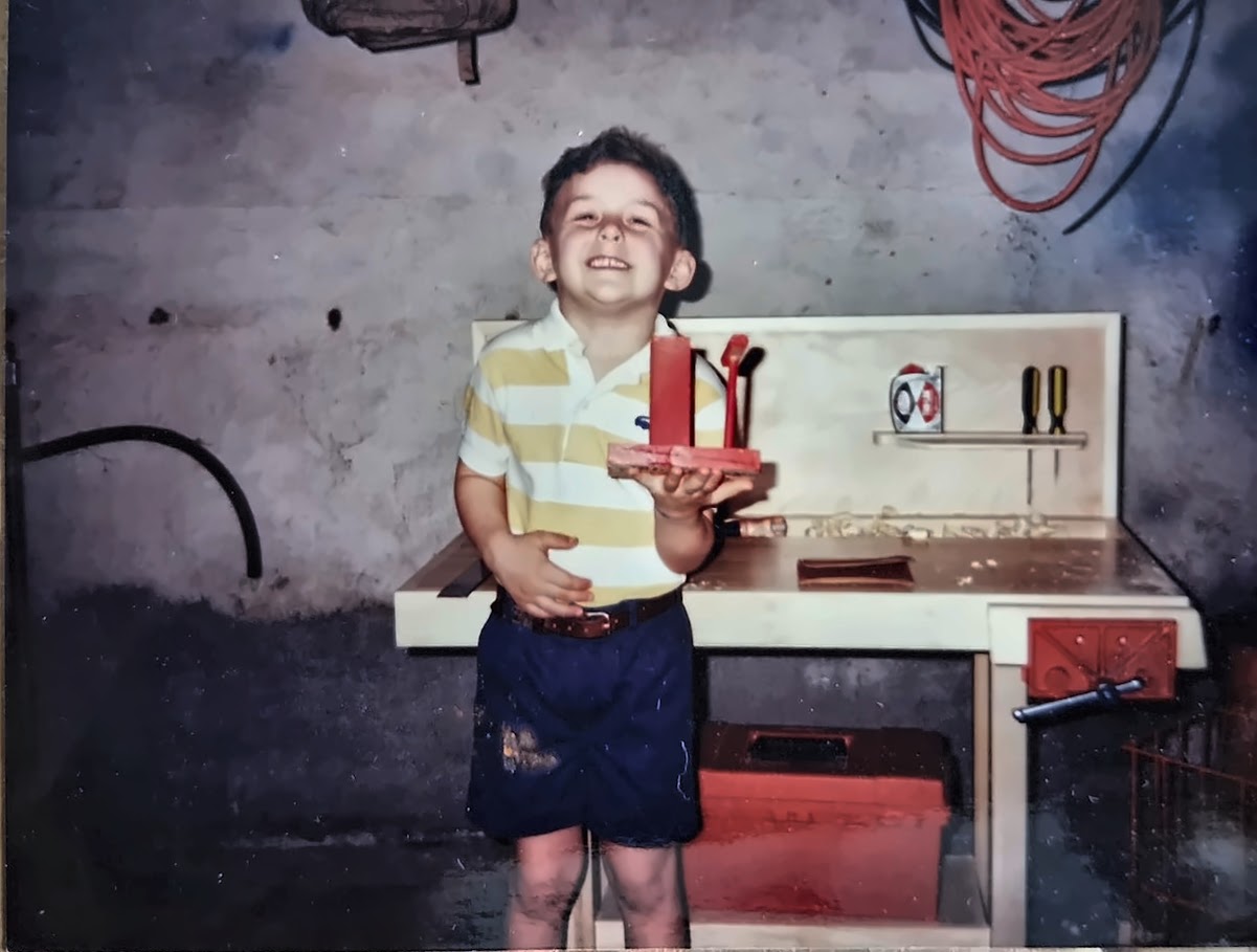Justin Sovine at about five, grinning in a striped shirt and holding something he’d just built, in front of the workbench his dad, Chris Sovine, built him