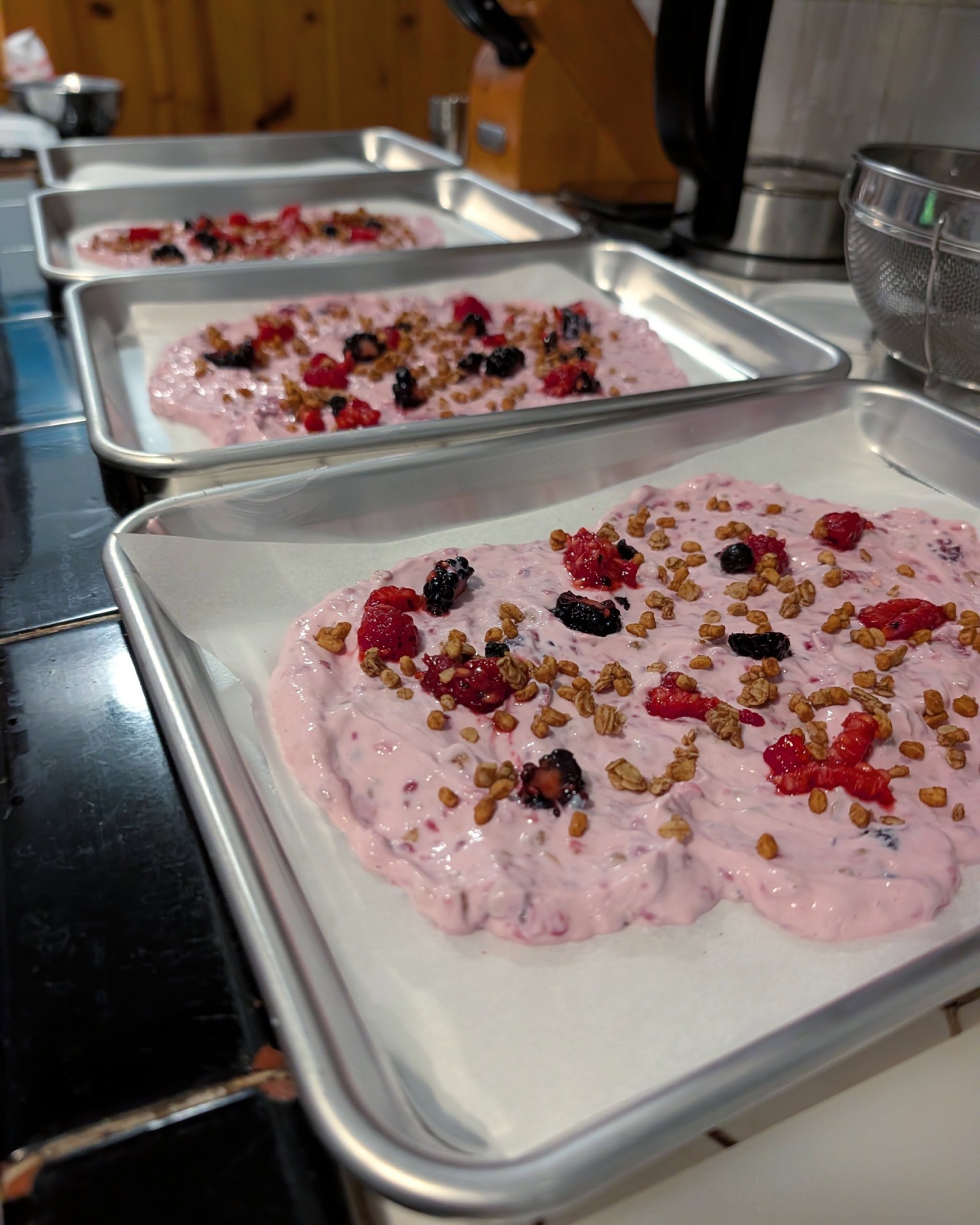 Three parchment-lined quarter sheets of pink frozen yogurt bark with berries and granola, before freezing