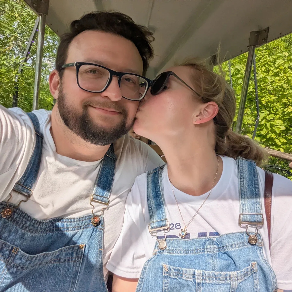 Justin Sovine smiling in glasses and denim overalls, getting a kiss on the cheek